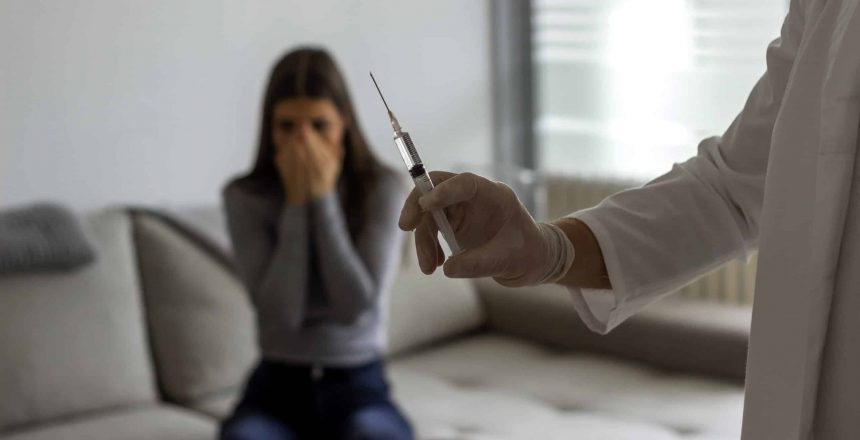 Male doctor or nurse is about to give shot of vaccine against virus to  afraid patient. Doctor giving nervous young girl an injection at home. A doctor preparing to give a upset young woman an vaccine